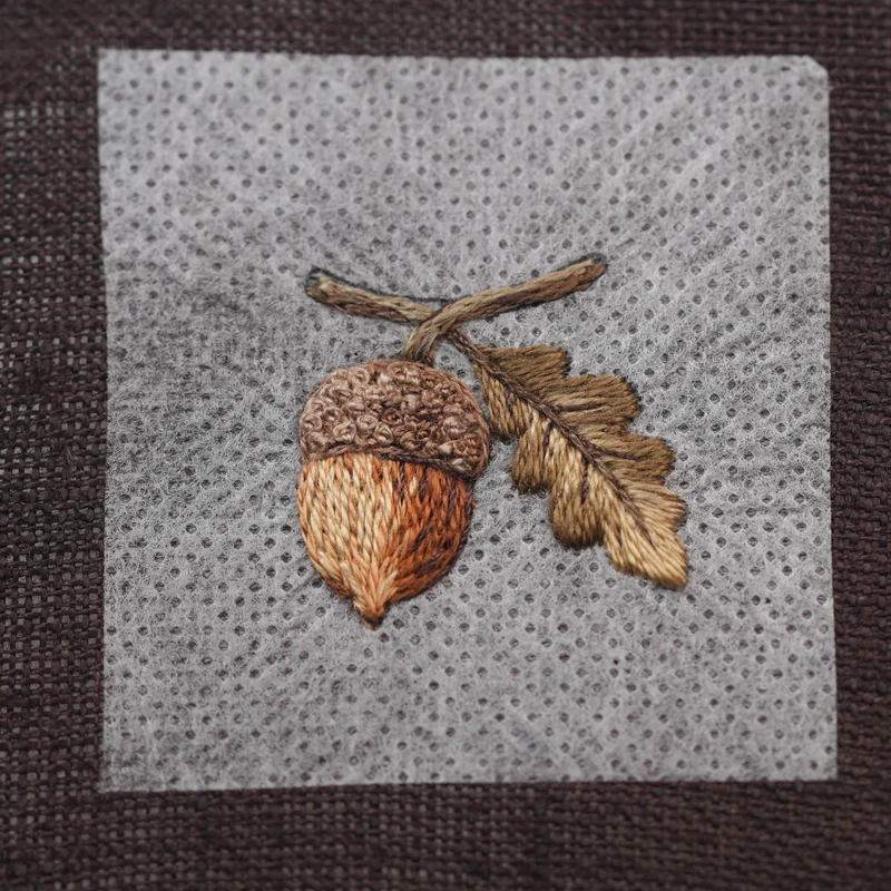 Using dark brown sewing thread, outline the lower edge of the branch and add the leaf veins. Add a few stitches beneath the acorn cap to create a subtle shadow, then add several stitches along the right side of the acorn for extra depth.